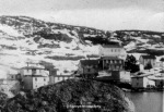 black and white photograph ofresettled community of bay du nord. A large boulder is seen in the foreground, white two-story houses in the middle ground and a snow-covered hillside in the background.